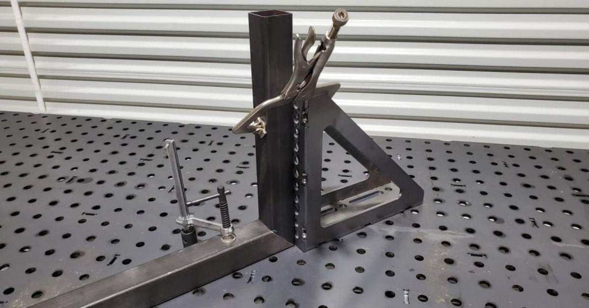Welding fixture setup on a perforated welding table using clamps and a 90-degree angle bracket to hold steel pieces in place